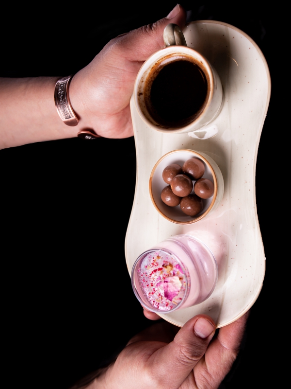 A Hand Holding A White Ceramic Tray Containing A Cup Of Espresso, Chocolate Treats, And A Small Pink Dessert.
